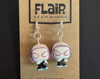 Puede incluir: Pendientes colgantes con figuras en miniatura de un superhéroe. Los pendientes están sujetos a ganchos plateados. Las figuras tienen una cara blanca, una máscara rosa y un atuendo negro y azul. Los pendientes se exhiben en una tarjeta marrón con el texto "FLAiR".