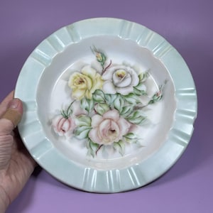 May include: A round ceramic ashtray featuring a pale mint green rim with integrated notches. The center of the white basin is decorated with a hand-painted floral arrangement consisting of yellow, white, and soft pink roses with green leaves.
