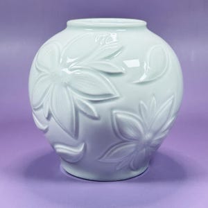 May include: A round, pale mint green ceramic vase featuring a glossy finish and a raised, embossed floral pattern across its surface. The vase has a wide, bulbous body that tapers toward a smooth, circular opening at the top.