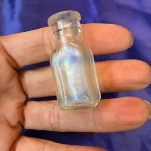 May include: A small, clear glass bottle with a rounded top and a rectangular body. The bottle has an iridescent sheen, reflecting multiple colours. The bottle is held in a hand.