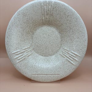 May include: A speckled, cream-coloured ceramic plate with a shallow bowl shape. The plate has etched lines on the rim and a central, recessed area. The plate is displayed on a clear stand. The plate is a decorative item.