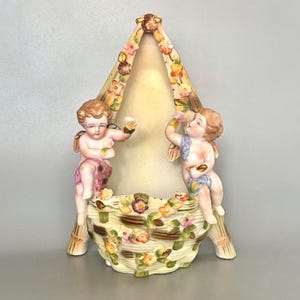 Puede incluir: Un marco de fotos de porcelana vintage con dos figuras de querubines alados sosteniendo flores. El marco tiene un fondo color crema y está decorado con detalles florales en rosa, amarillo y marrón. El marco mide aproximadamente 25 cm de alto.