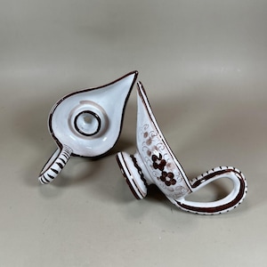 Może przedstawiać: Dwie białe ceramiczne lampy oliwne z brązowymi wykończeniami i kwiatowymi wzorami. Jedna lampa stoi pionowo, druga leży na boku. Lampy mają uchwyt i wylew. Lampy są prawdopodobnie zabytkowe lub antyczne.