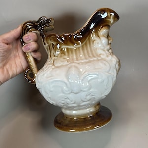May include: A decorative ceramic pitcher with a brown and white color scheme. The pitcher features a detailed handle with a dragon-like design and a face sculpted on the side. The base is a pedestal design.