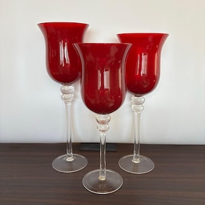 May include: Three tall glass candle holders featuring deep red, tulip-shaped bowls atop clear, slender stems with a decorative spherical accent near the base. Each piece stands on a circular, transparent glass foot, creating a uniform set.