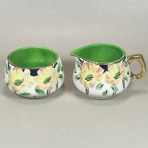 May include: A ceramic creamer and sugar bowl set featuring a white base with hand-painted yellow flowers, green leaves, and black accents. Both pieces have a vibrant green interior and a metallic gold rim. The creamer includes a gold-toned handle.