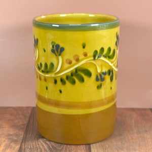 以下が含まれることがあります： 緑色の縁と茶色のベースが付いた、円筒形の黄色のセラミック花瓶。花瓶は、緑の葉と青のアクセントが付いた、隆起した白い花の模様が特徴です。茶色とオレンジ色の点が花の模様の周りに散らばっています。