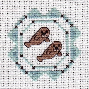 May include: Cross-stitch pattern of two brown seals swimming in a blue and white frame.