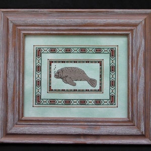 May include: A framed cross-stitch embroidery featuring a brown manatee swimming in a turquoise blue water with bubbles. The embroidery is surrounded by a multi-colored border.