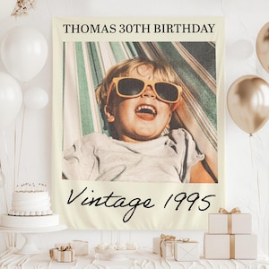 May include: A cream-colored tapestry with a vintage photo of a child wearing sunglasses. The text "THOMAS 30TH BIRTHDAY" is at the top, and "Vintage 1995" is written below the photo. The tapestry is part of a birthday party setup.