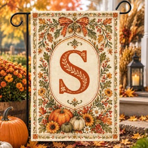 May include: A rectangular garden flag featuring a large orange letter S centered within an oval frame. The design is decorated with autumn leaves, berries, sunflowers, and pumpkins. A plaid bow sits at the top, and the background is cream-colored.