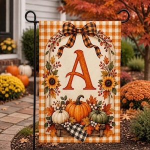 May include: A rectangular garden flag featuring an orange and white buffalo check border. The center displays a large orange letter A framed by sunflowers, autumn leaves, berries, and a plaid bow. Three pumpkins sit at the base on a wooden crate.
