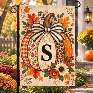May include: A rectangular garden flag featuring a large central pumpkin with a black letter S. The pumpkin has sections with orange gingham, floral, and leopard patterns, topped with a black and white striped bow, surrounded by autumn leaves.