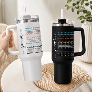 May include: Two tall stainless steel tumblers with handles and clear lids with straws. One tumbler is white and the other is black. Both feature the name Samuel in cursive script next to a vertical column of colorful embroidered stitch patterns.
