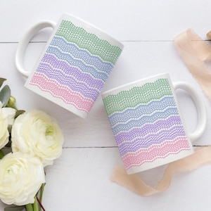 May include: Two white ceramic mugs with a colorful lace design in green, blue, purple, and pink. The mugs are placed on a white wooden surface, next to white flowers and a beige ribbon.