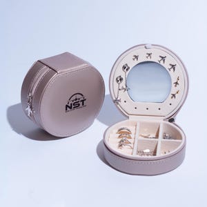 May include: A round, light brown, faux leather jewelry box with a mirror and compartments for rings, earrings, and other small jewelry. The box has a zipper closure and a logo that says "NST - New Style Traveler".