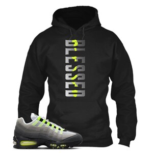 Op de afbeelding: Zwarte hoodie met het woord "BLESSED" in een verticaal ontwerp. De hoodie wordt gecombineerd met een grijze en zwarte sneaker met neon groene accenten.