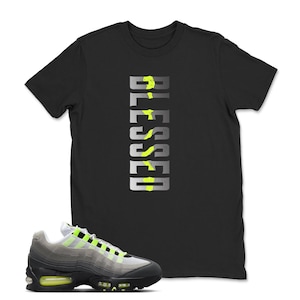 Op de afbeelding: Zwart T-shirt met het woord "BLESSED" in een verticaal ontwerp met neon groene accenten. Een paar grijze en zwarte sneakers met neon groene details staan op de voorgrond.