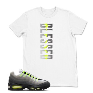 Op de afbeelding: Wit T-shirt met het woord "BLESSED" in een verticaal ontwerp, met neon groene accenten. Een paar grijze, zwarte en neon groene sportschoenen staan op de voorgrond, met een zwarte zool en neon groene accenten.