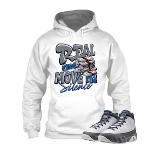 May include: White hoodie with the text "REAL ONCE MOVE IN SILENCE" in gray and blue. The graphic includes a stack of money. The image also shows a pair of white and gray sneakers with blue accents.
