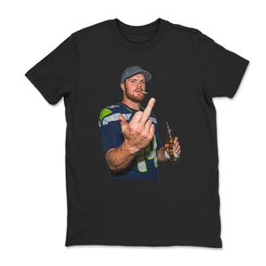 May include: Black t-shirt featuring a person wearing a football jersey, a cap, and holding a beer bottle and cigarette. The person is giving the middle finger. The jersey is blue, green, and white.