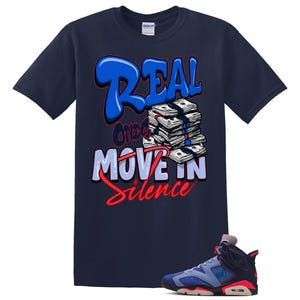 May include: A navy blue short-sleeve t-shirt featuring a graphic print of a stack of cash and the text "REAL ONES MOVE IN SILENCE" in blue, white, and red lettering, accompanied by a single high-top sneaker in matching navy, blue, and red colors.