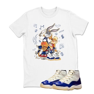 May include: White t-shirt featuring a cartoon graphic of Bugs Bunny and Lola Bunny. The graphic includes money, diamonds, and hearts. A pair of white and blue sneakers are in the foreground.