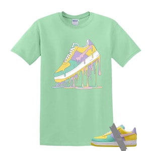 May include: A light green t-shirt featuring a colorful graphic of a sneaker with dripping paint in yellow, purple, and blue. A matching sneaker is taped below the shirt.