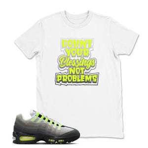 May include: White t-shirt with the text "Count Your Blessings Not Problems" in neon green and gray. A pair of gray, black, and neon green sneakers is in the foreground.