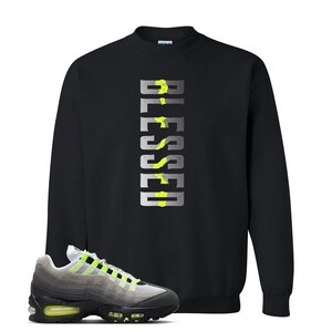 Op de afbeelding: Zwart sweatshirt met het woord "BLESSED" in een verticaal, zilver en neon groen ontwerp. Een paar grijze, zwarte en neon groene sneakers staan op de voorgrond.
