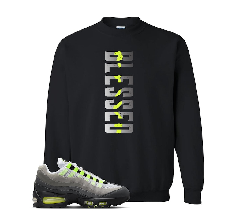 Op de afbeelding: Zwart sweatshirt met het woord "BLESSED" in een verticaal, zilver en neon groen ontwerp. Een paar grijze, zwarte en neon groene sneakers staan op de voorgrond.