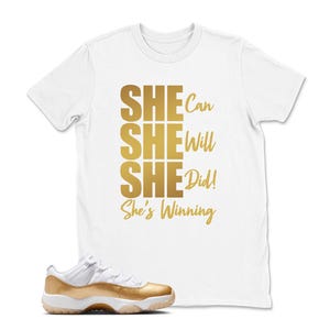 May include: White t-shirt with gold text that reads "SHE Can, SHE Will, SHE Did! She's Winning." A gold and white sneaker is in the foreground. The t-shirt is a crew neck style.