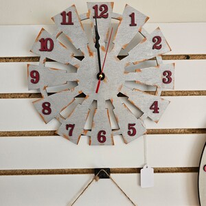 Puede incluir: Un reloj blanco y plateado en forma de molino de viento con n&uacute;meros rojos. El reloj est&aacute; colgado en una pared blanca con listones de madera marr&oacute;n.