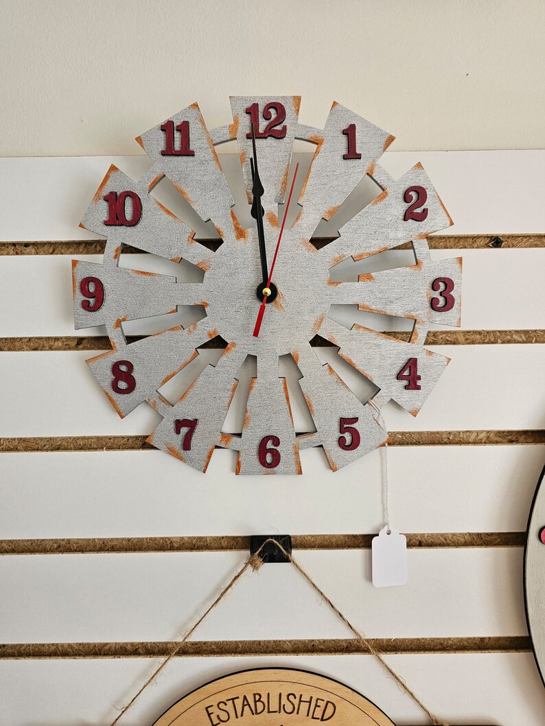 Puede incluir: Un reloj blanco y plateado en forma de molino de viento con n&uacute;meros rojos. El reloj est&aacute; colgado en una pared blanca con listones de madera marr&oacute;n.