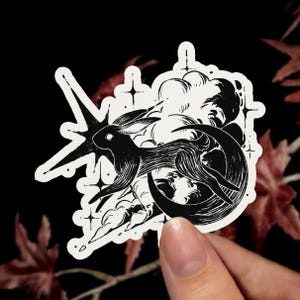 Peut inclure: Un autocollant découpé en noir et blanc représentant la silhouette d'un lapin sautant à travers un croissant de lune, entouré de nuages stylisés et d'une grande étoile à quatre branches avec des accents d'étoiles plus petites.