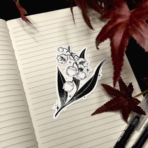 May include: A black and white sticker featuring a detailed illustration of a lily of the valley plant with bell-shaped flowers and pointed leaves, accented by small four-pointed star shapes, displayed on a lined notebook page.