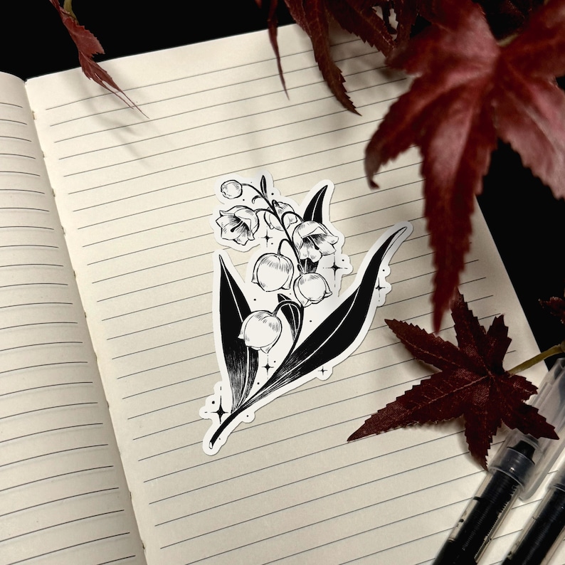 May include: A black and white sticker featuring a detailed illustration of a lily of the valley plant with bell-shaped flowers and pointed leaves, accented by small four-pointed star shapes, displayed on a lined notebook page.