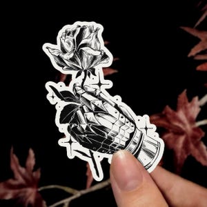 Peut inclure: Un autocollant en noir et blanc représentant une illustration détaillée d'un gantelet métallique blindé tenant une rose unique. Le motif comprend de petites étoiles à quatre branches entourant la main et la fleur, dans un style à l'encre contrasté.