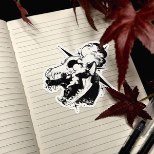 K&ouml;nnte beinhalten: Ein schwarz-wei&szlig;er Aufkleber mit einer stilisierten Illustration eines Wolfskopfes mit Totenkopf-&Uuml;berlagerung und wirbelnden Raucheffekten. Das Design enth&auml;lt scharfe geometrische Linien und kleine Sternakzente in kontrastreicher Tinte.