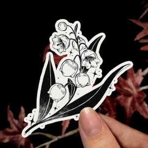 May include: A black and white sticker featuring a detailed line art illustration of a lily of the valley plant. The design includes several bell-shaped flowers and broad, solid black leaves, accented with small star shapes and dots.