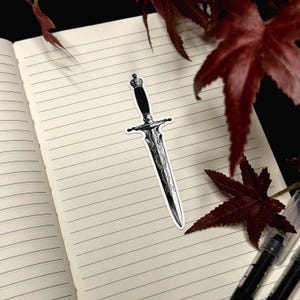May include: A black and white sticker of a dagger with an ornate, patterned blade and a dark handle. The dagger features a pointed tip, a crossguard, and a pommel at the top. The design is rendered in high-contrast ink style with fine line details.