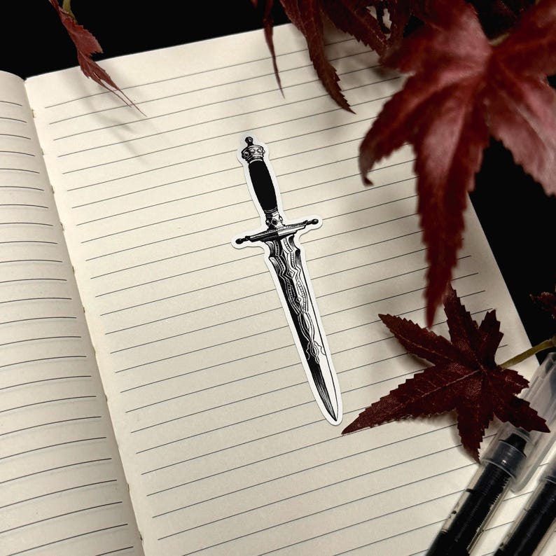 May include: A black and white sticker of a dagger with an ornate, patterned blade and a dark handle. The dagger features a pointed tip, a crossguard, and a pommel at the top. The design is rendered in high-contrast ink style with fine line details.