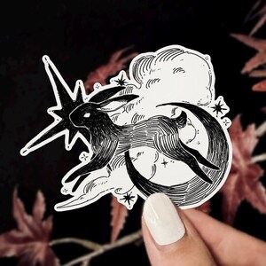 Puede incluir: Pegatina en blanco y negro que representa un conejo saltando sobre una luna creciente, con una estrella fugaz y nubes. La ilustraci&oacute;n tiene un tema celestial caprichoso, con peque&ntilde;as estrellas dispersas. La pegatina est&aacute; sujeta por una persona.