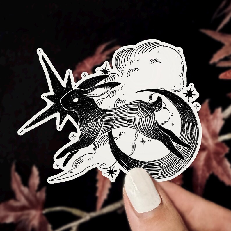 Puede incluir: Pegatina en blanco y negro que representa un conejo saltando sobre una luna creciente, con una estrella fugaz y nubes. La ilustraci&oacute;n tiene un tema celestial caprichoso, con peque&ntilde;as estrellas dispersas. La pegatina est&aacute; sujeta por una persona.