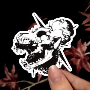 K&ouml;nnte beinhalten: Ein schwarz-wei&szlig;er Aufkleber mit einer stilisierten Illustration eines Wolfskopfes, der einen Sch&auml;del als Kopfschmuck tr&auml;gt. Das Design zeigt wirbelnden Rauch, spitze Z&auml;hne und kleine vierzackige Sterne in kontrastreichen Tuschelinien.