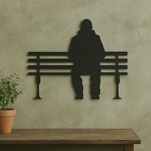 Puede incluir: Arte de pared de metal negro que representa a una persona sentada en un banco de parque. La silueta está montada en una pared texturizada de color verde claro. Una planta en maceta está sobre una superficie de madera.