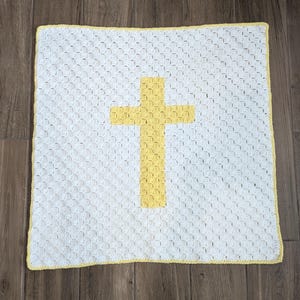 May include: White crocheted baby blanket with a yellow cross in the center. The blanket has a yellow border.