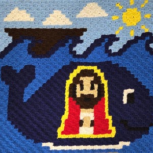 May include: A handmade crochet blanket featuring a blue whale with a figure inside, set against a blue ocean, white clouds, and a yellow sun. The blanket is made with various shades of blue, brown, yellow, red, and white yarn.