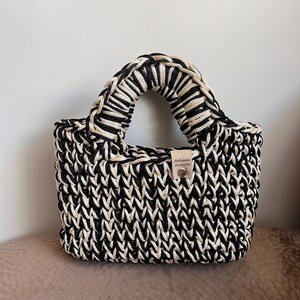 May include: A small, hand-crocheted tote bag in black and cream. The bag features a woven pattern and a sturdy handle. A small leather tag with the text "Artisan Avenue" is attached to the side.