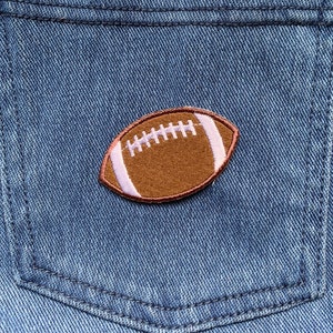 Può includere: Un toppa di cuoio marrone a forma di pallone da football con cuciture bianche cucita su una tasca di jeans blu.
